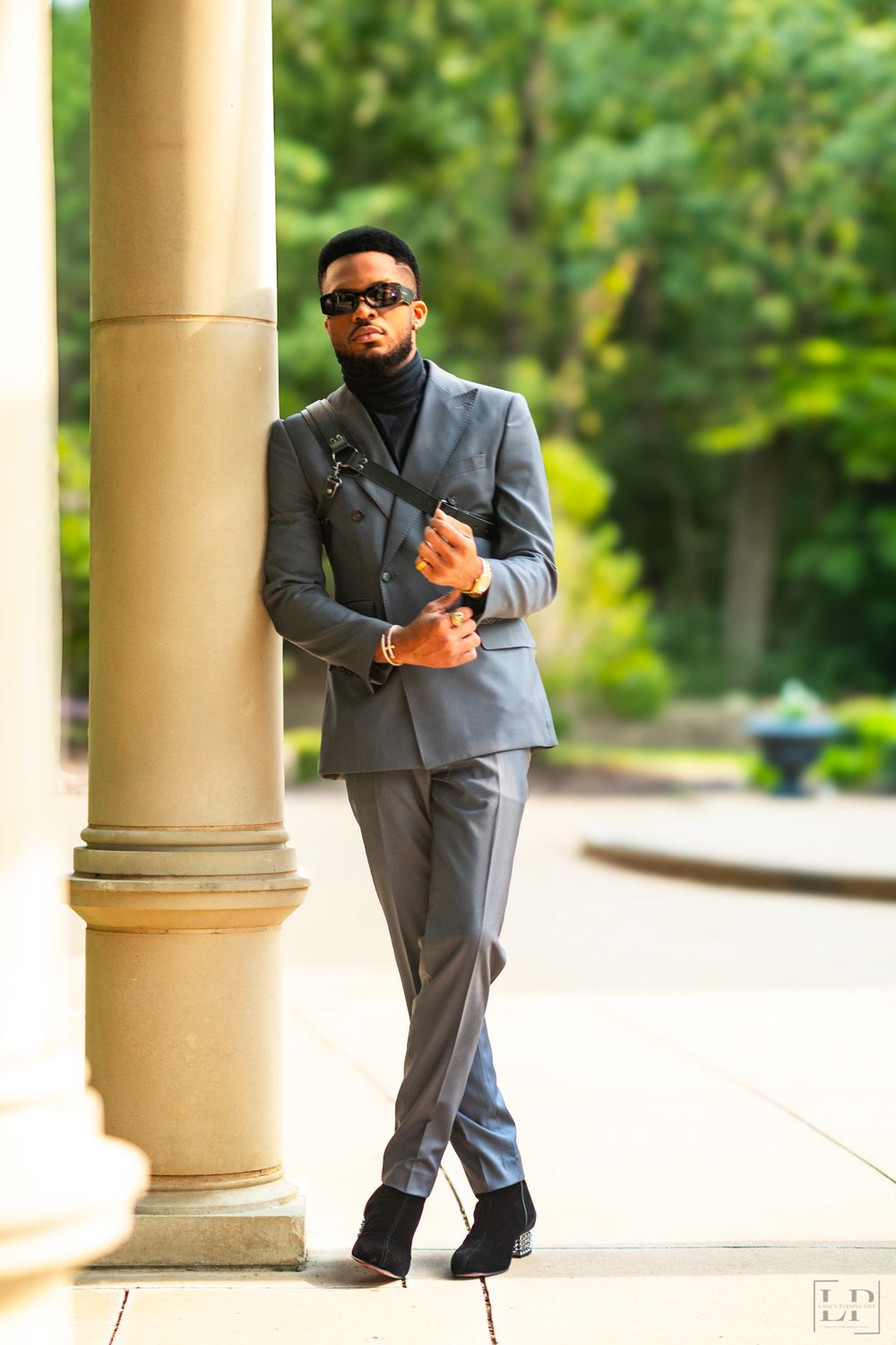 A person in a grey suit stands between beige columns in an outdoor architectural setting.