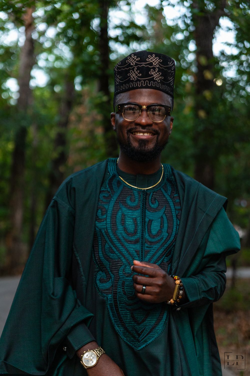 A person wearing a traditional green embroidered agbada poses outdoors among trees.