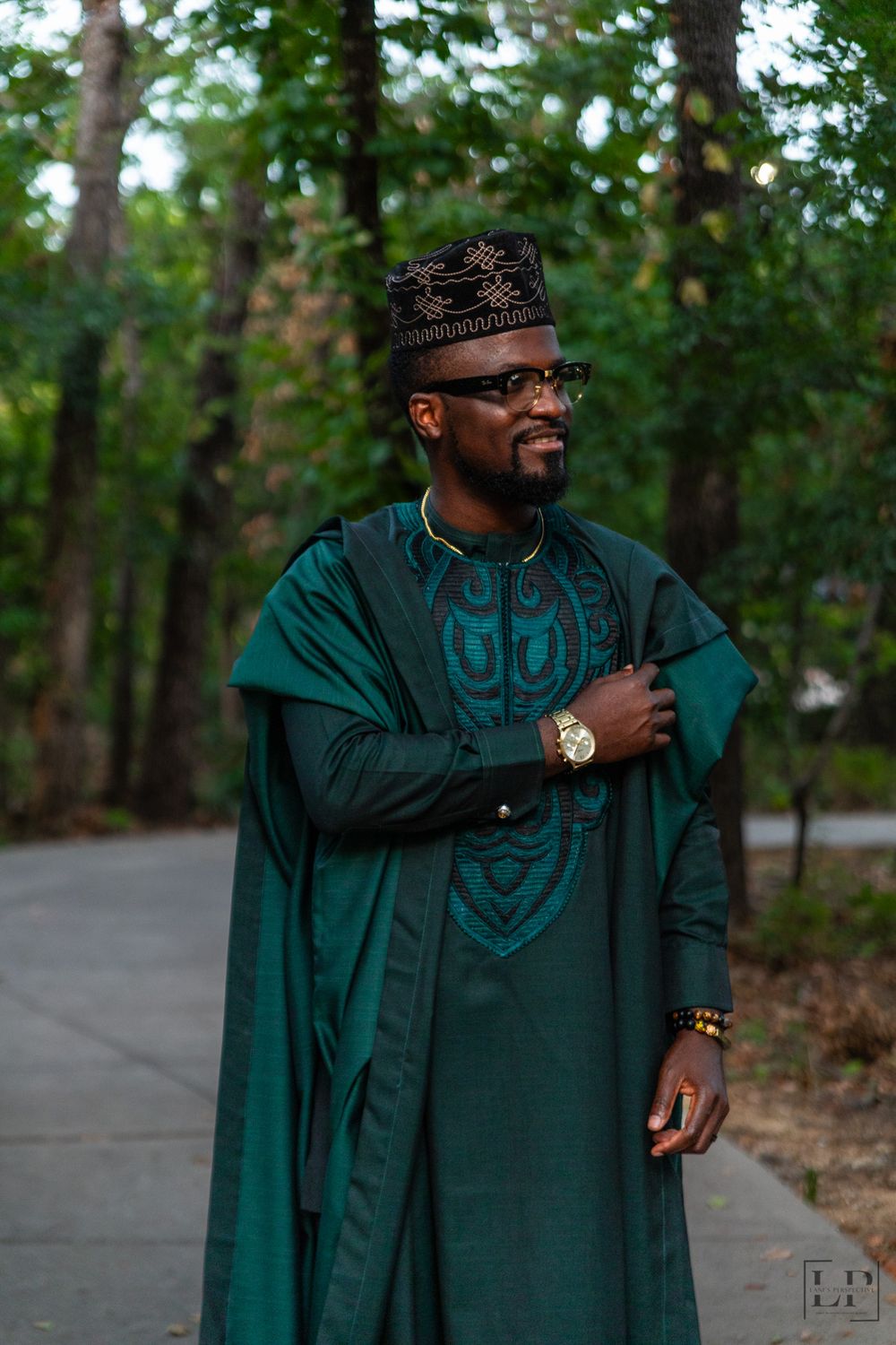 A person in an elegant dark green traditional outfit smiles while standing in a wooded outdoor setting.