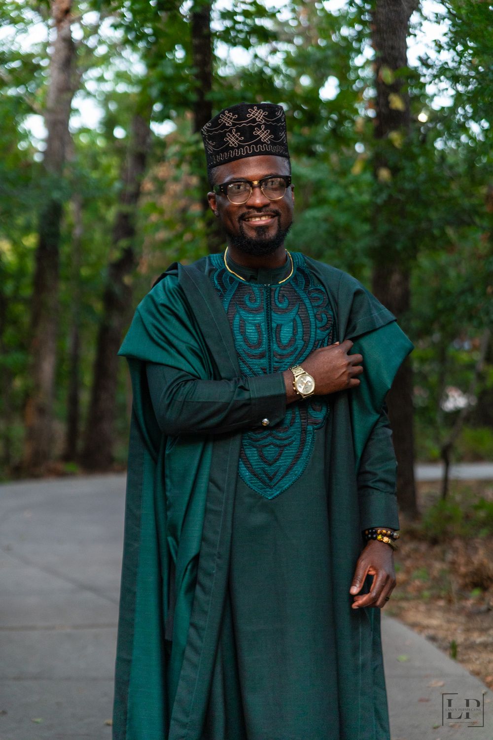 A person wearing a dark green traditional garment and cap poses confidently in a natural setting.