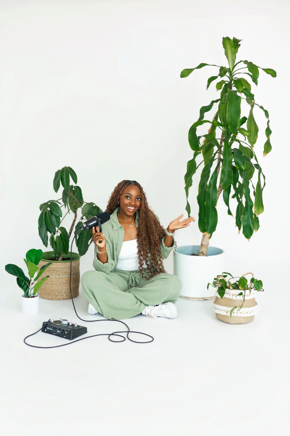 Person in mint green sweater sitting among tropical houseplants against white background.