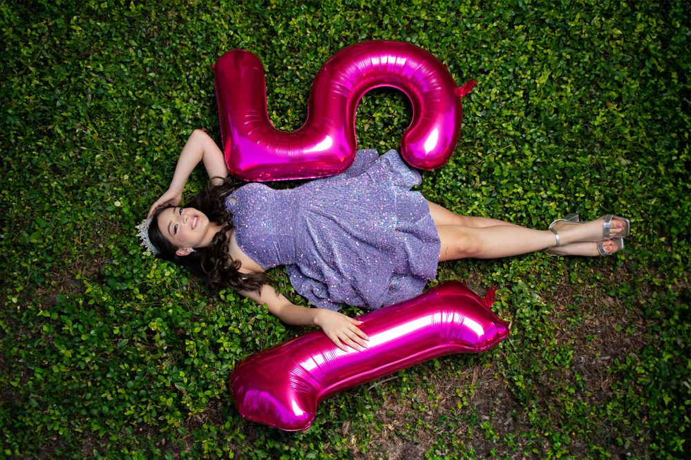 Purple sparkly dress contrasts with pink number balloon decoration on green grass.