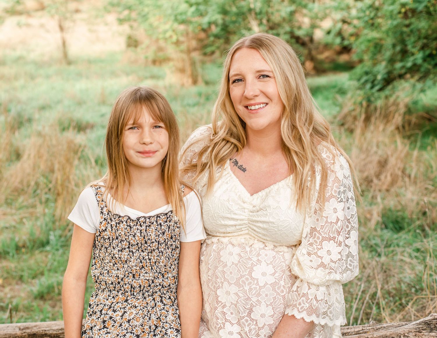 Two people in coordinating floral and lace dresses pose together outdoors in a sunlit natural setting.