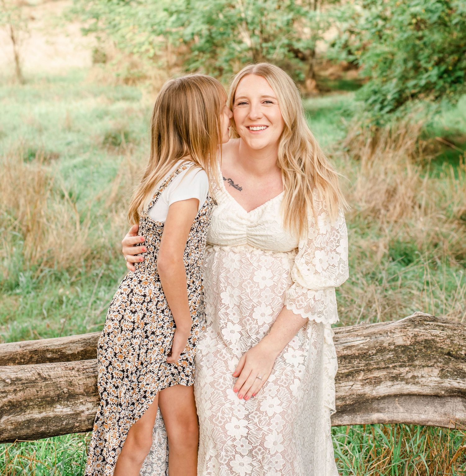 Two people in floral dresses share a joyful moment together while sitting on a fallen tree trunk in a sunny meadow.