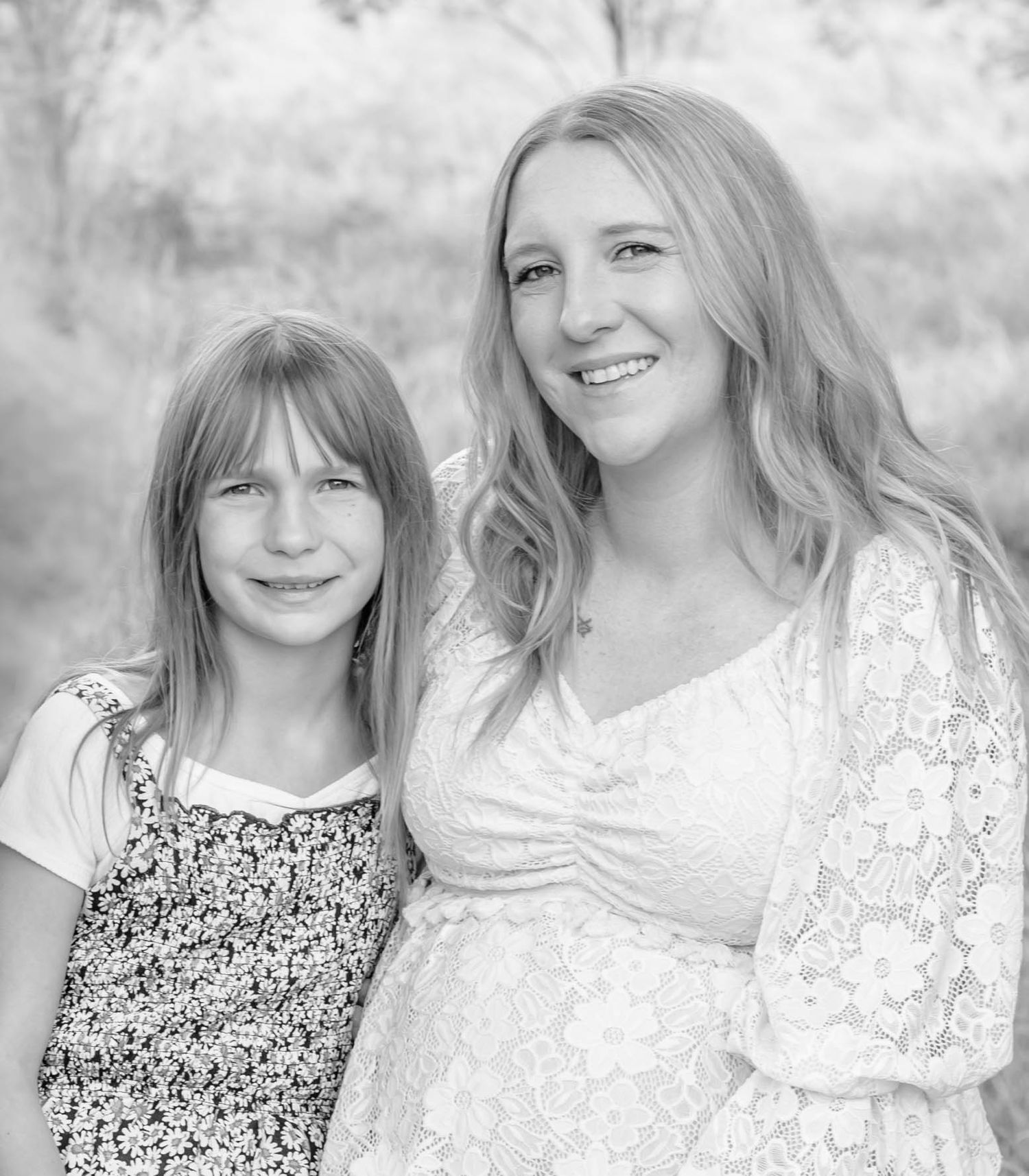 Black and white portrait of a mother and daughter smiling together outdoors in matching floral dresses.