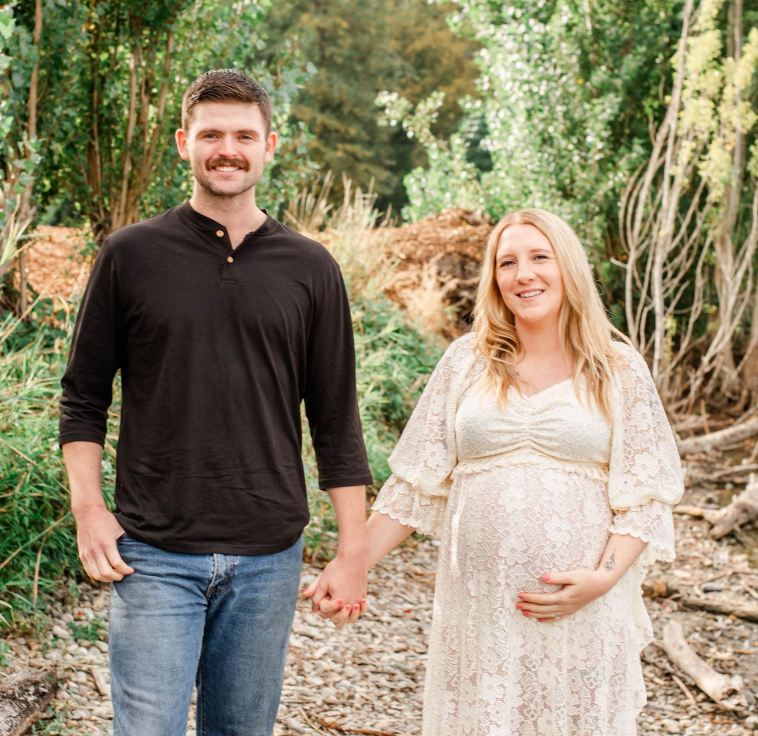 A couple holding hands in a wooded outdoor setting wearing casual attire and sharing intimate moments together.