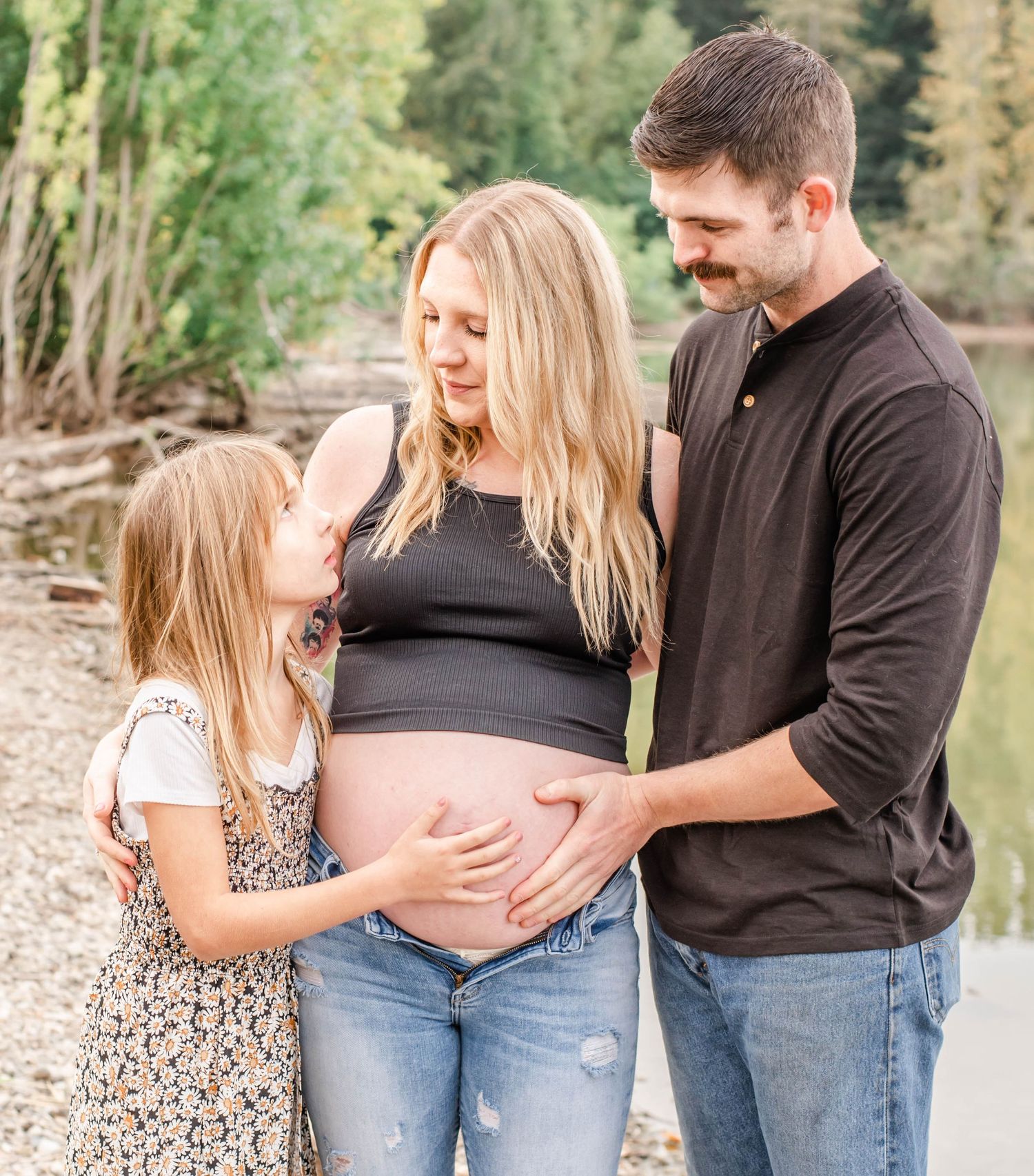 Family maternity photo session outdoors with daughter lovingly touching pregnant mother's belly.