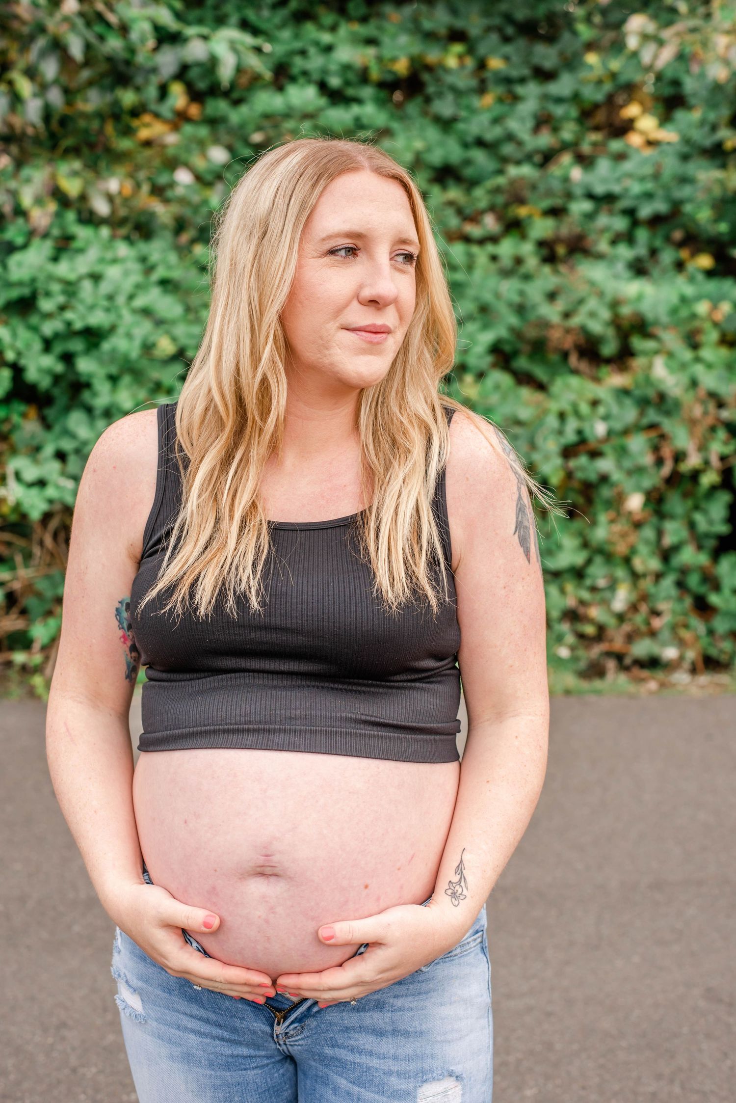 A pregnant person in a black tank top and jeans stands outdoors showing their growing belly against a green foliage backdrop.