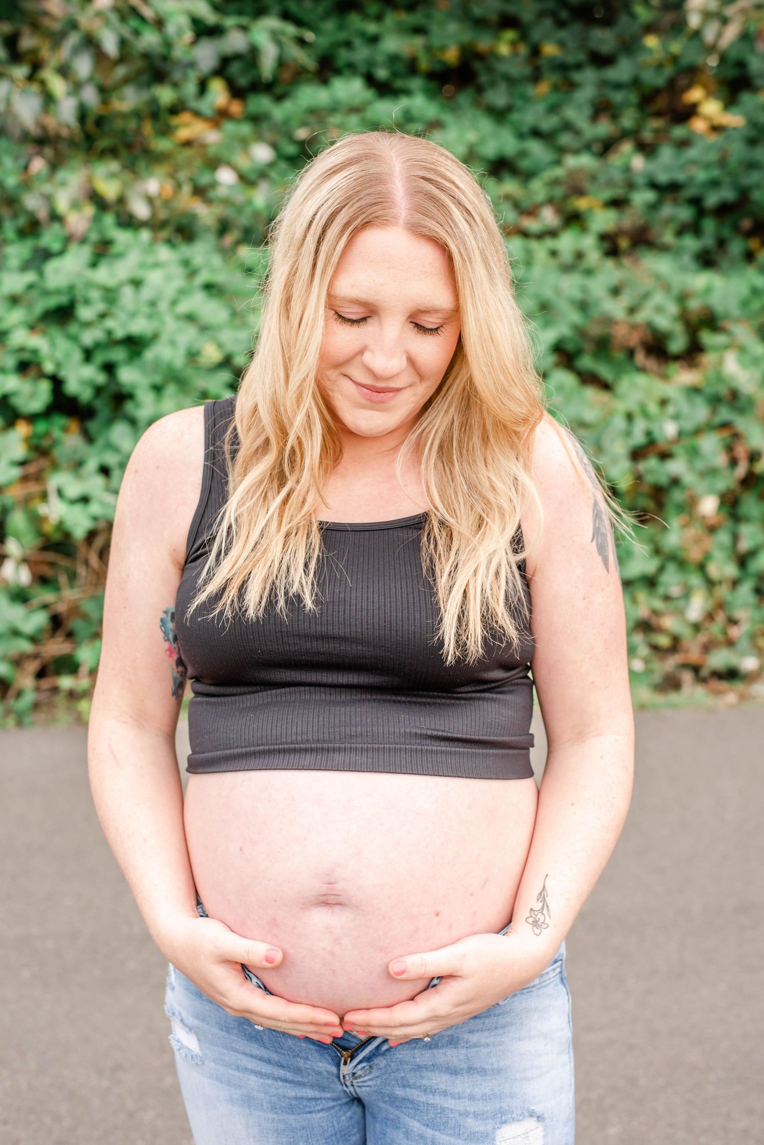A pregnant woman in a black crop top and jeans stands against a green leafy backdrop.
