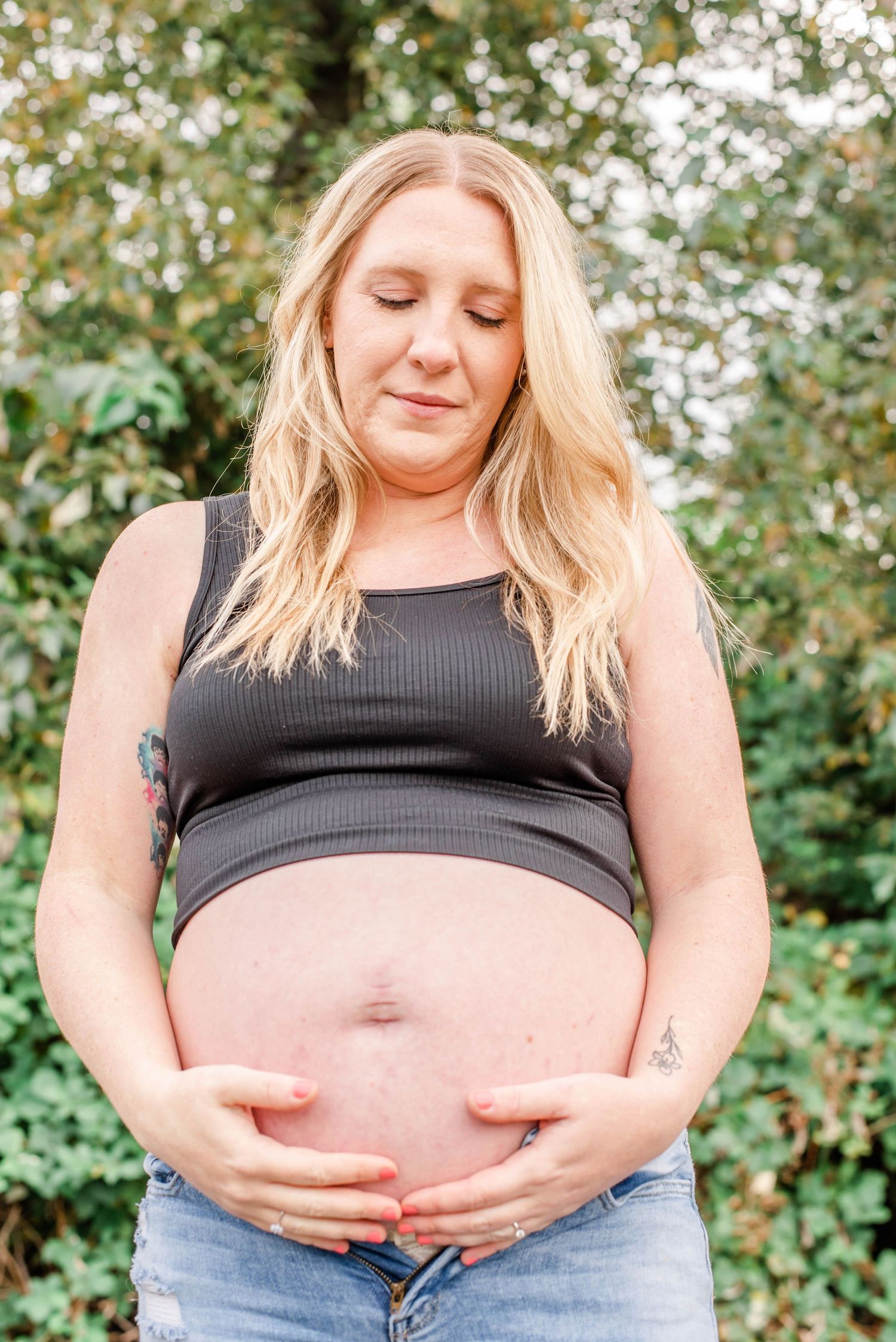 A pregnant woman in a black crop top and jeans stands against a green leafy backdrop.