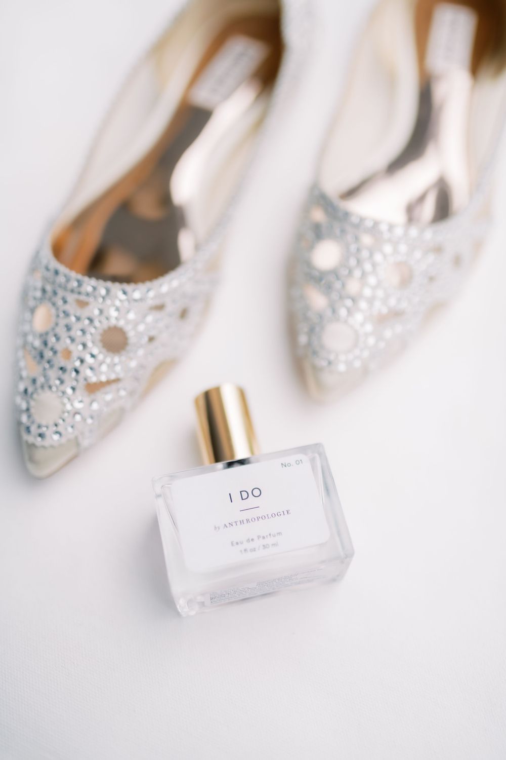 Wedding detail shot of sparkly bridal shoes and 'I DO' perfume bottle on white background.