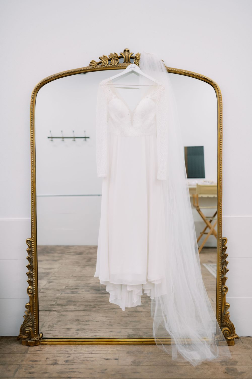Elegant white wedding dress hanging on ornate gold mirror frame with flowing veil in a minimalist room setting.
