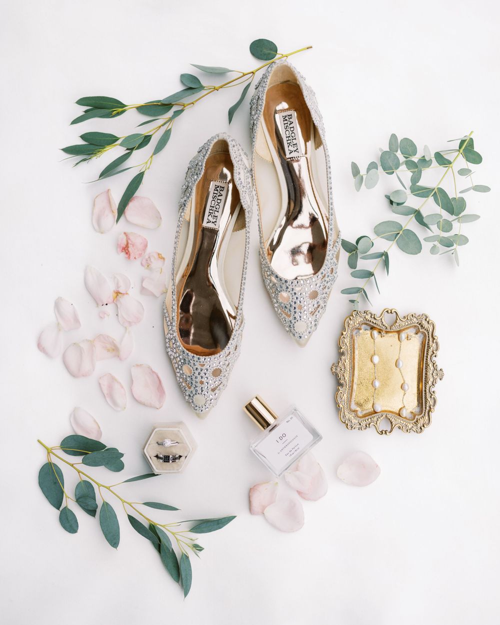 Sparkly bridal flats surrounded by eucalyptus leaves and rose petals on white background.