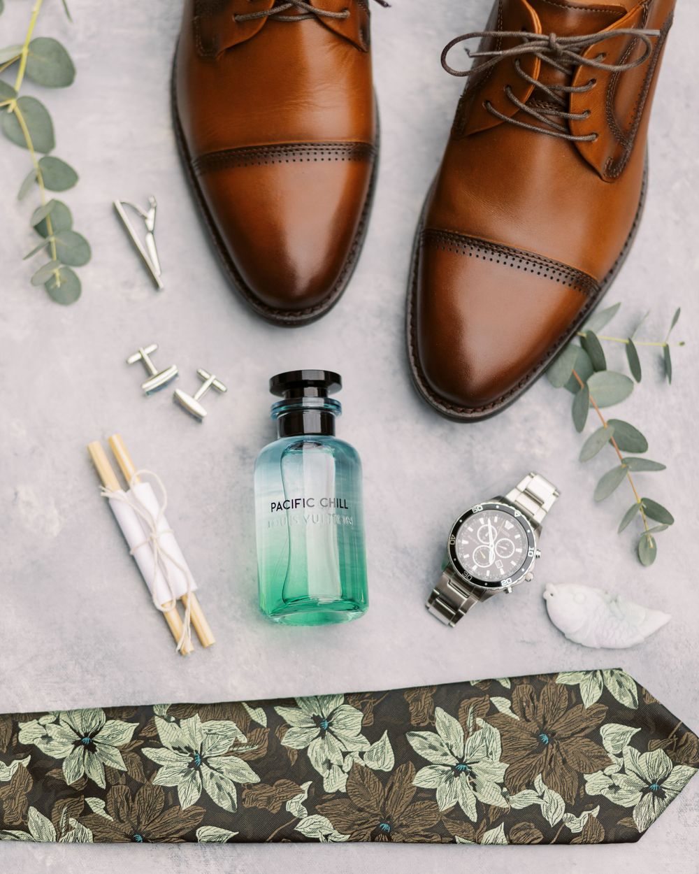 Wedding accessories including brown leather shoes, floral tie, cologne, and watch on gray background.