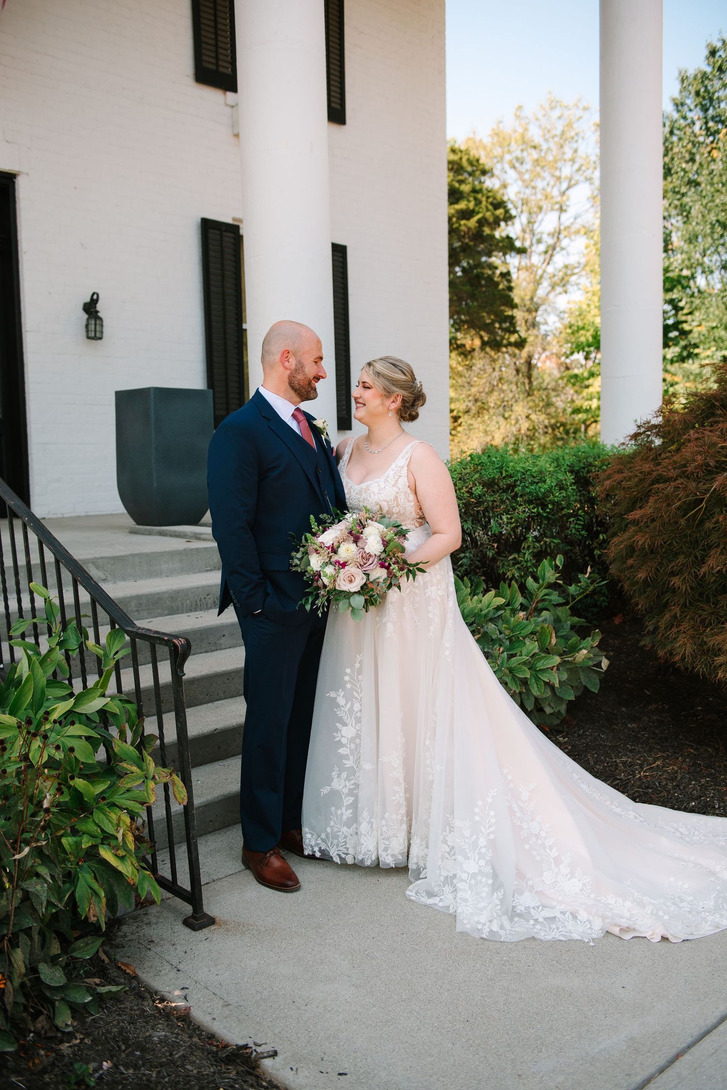 Newlyweds pose together on white pillared porch, bride in lace gown holding pink bouquet.