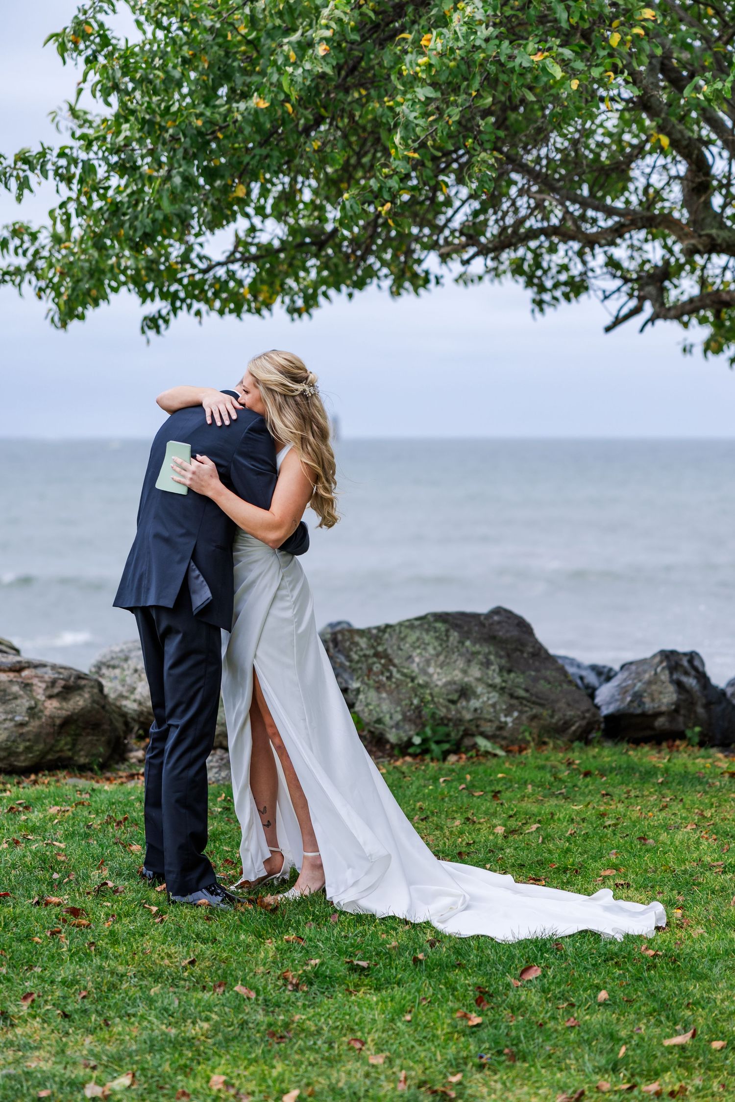 Romantic embrace between newlyweds on a grassy oceanfront with rocks and waves in background.