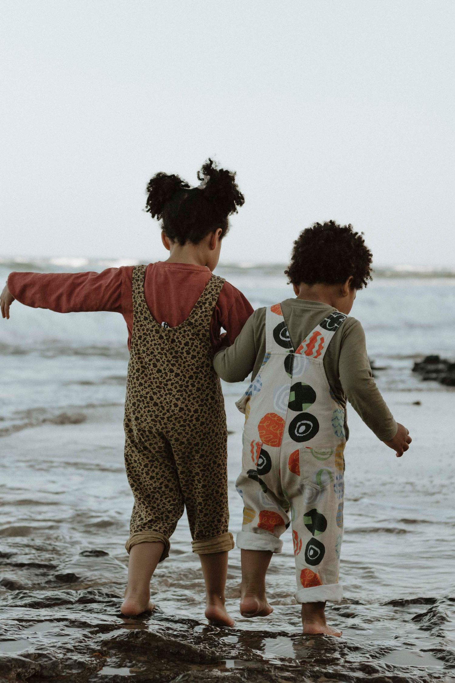 Two children in patterned overalls playing at the edge of ocean water.