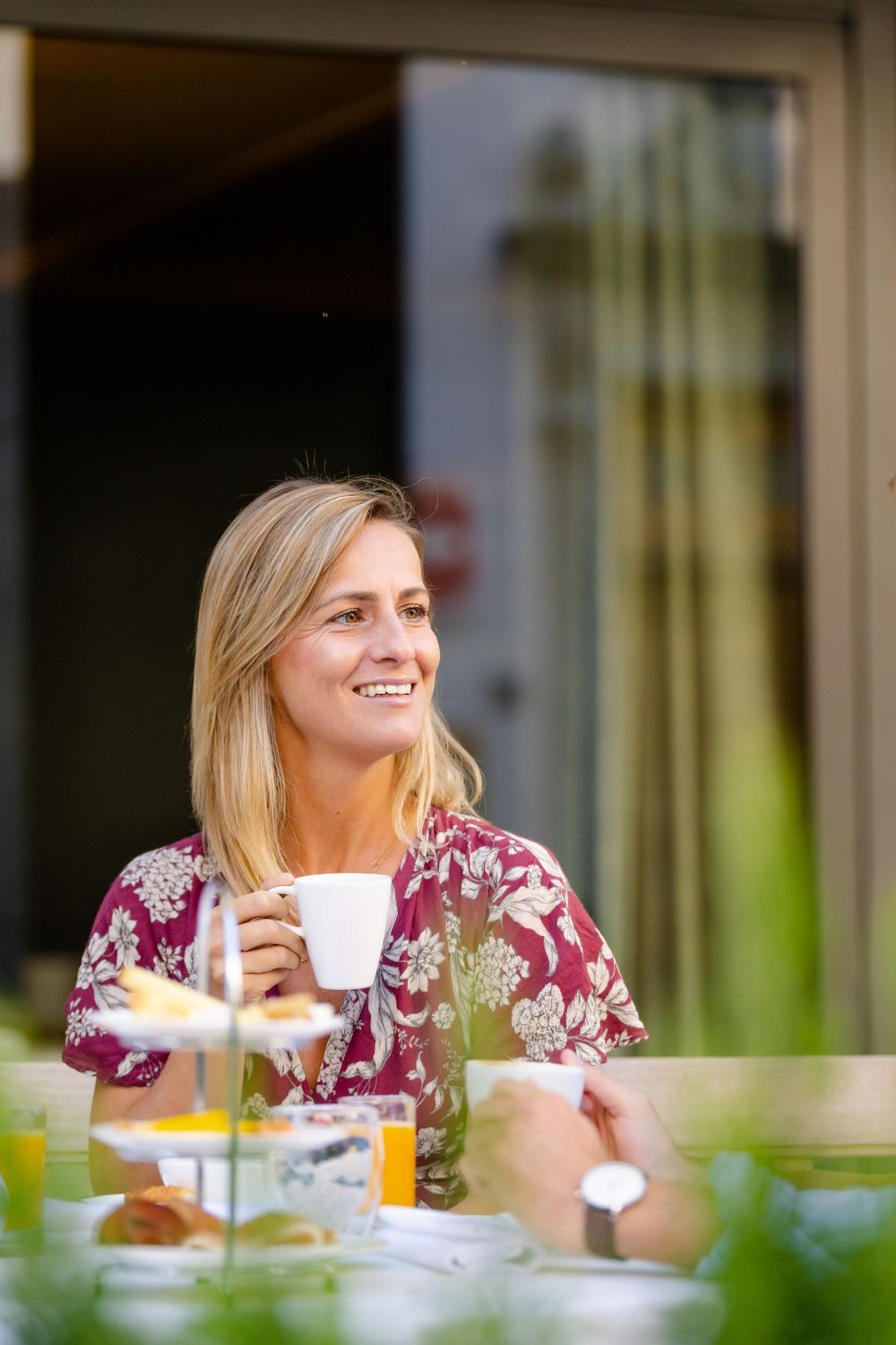 A woman in a floral dress smiles while holding a coffee cup at an outdoor cafe.