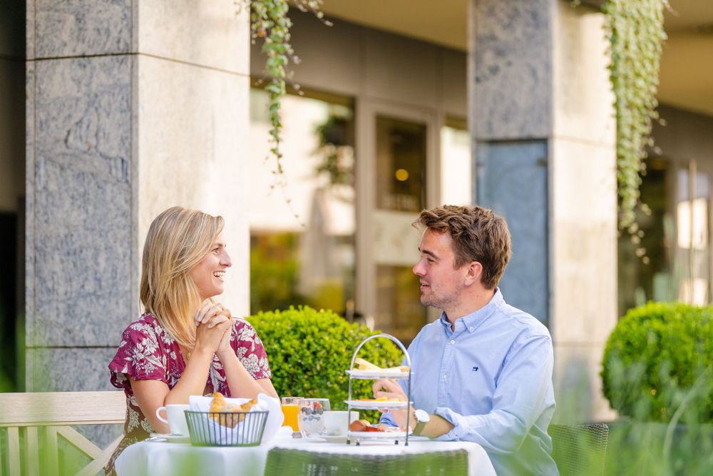 A couple enjoys an elegant afternoon tea service with pastries at an outdoor dining terrace surrounded by stone columns.