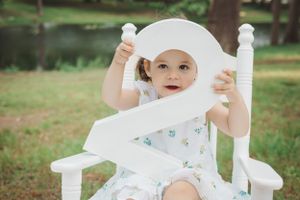 A toddler in a white floral dress holds up a number two while sitting in a white chair outdoors.