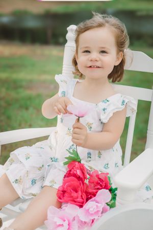 A toddler in a floral dress sits in a white chair with pink flowers outdoors.
