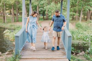 A family of three holds hands while walking across a blue wooden bridge in a park.