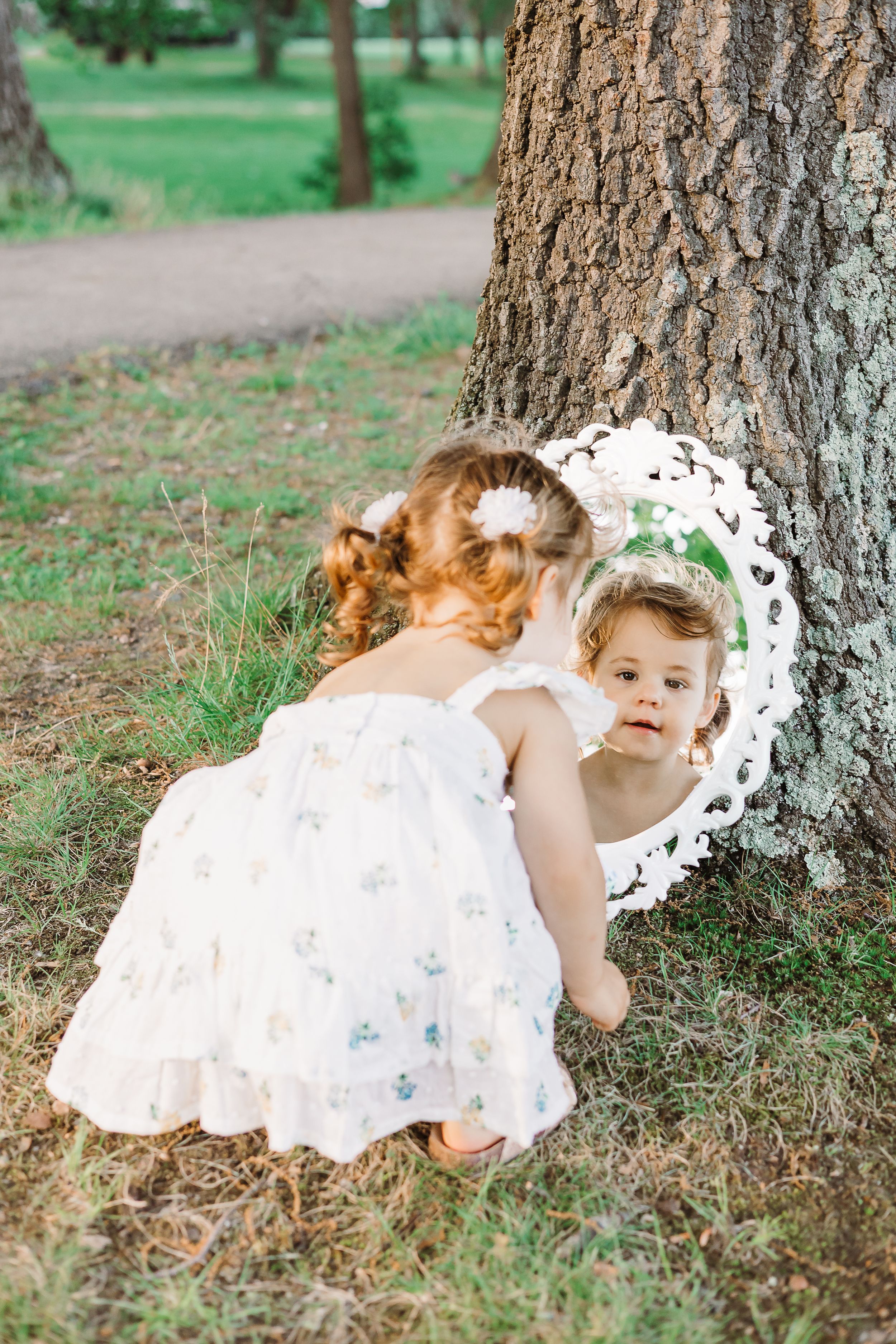 A child in a white dress looks into an ornate mirror propped against a tree trunk.