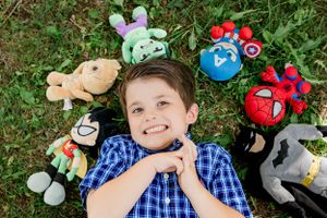 Plush toys and stuffed animals surround someone lying in green grass looking upward.