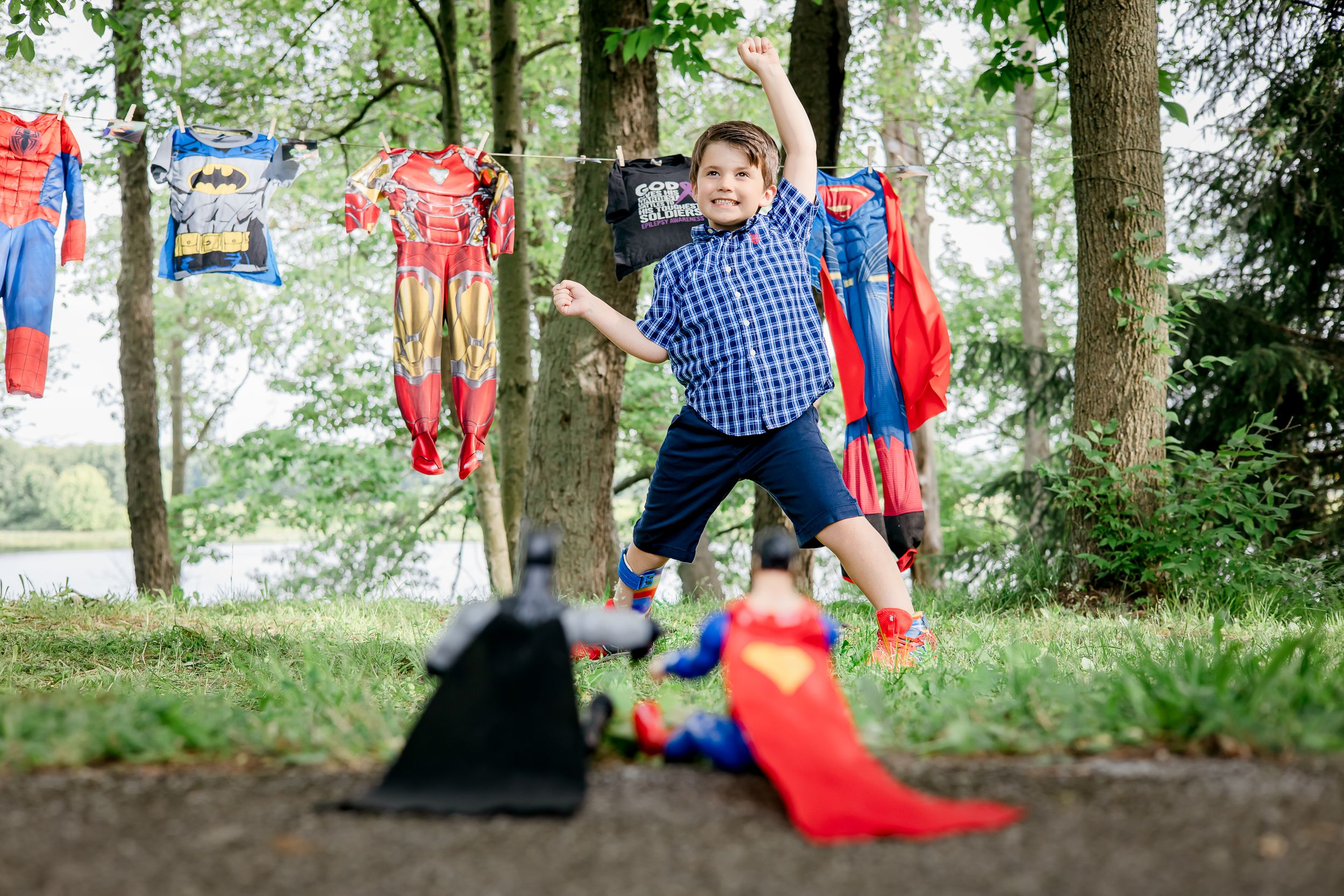 Superhero costumes hang from trees in an outdoor setting while someone jumps playfully.
