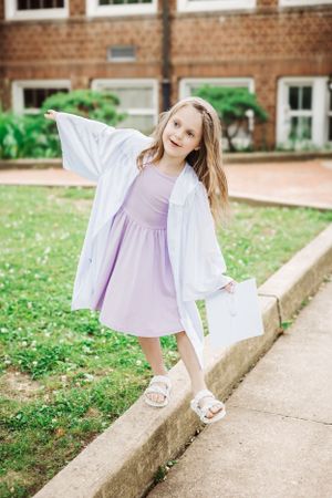 White graduation gown and lavender dress worn while balancing on concrete edge by brick building.