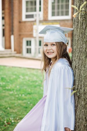 White cap and gown worn with purple dress while leaning against tree by brick building.
