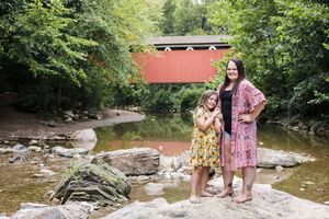 A family explores a scenic creek beneath a rustic red covered bridge on a sunny summer day.