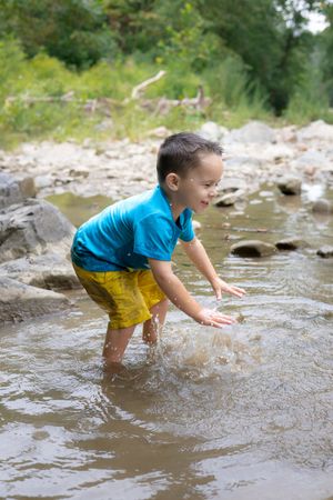 A young child wearing a blue shirt and yellow shorts plays in a shallow stream on a sunny summer day.