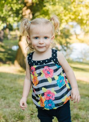 Toddler in floral striped tank top smiles brightly in sunny outdoor setting.