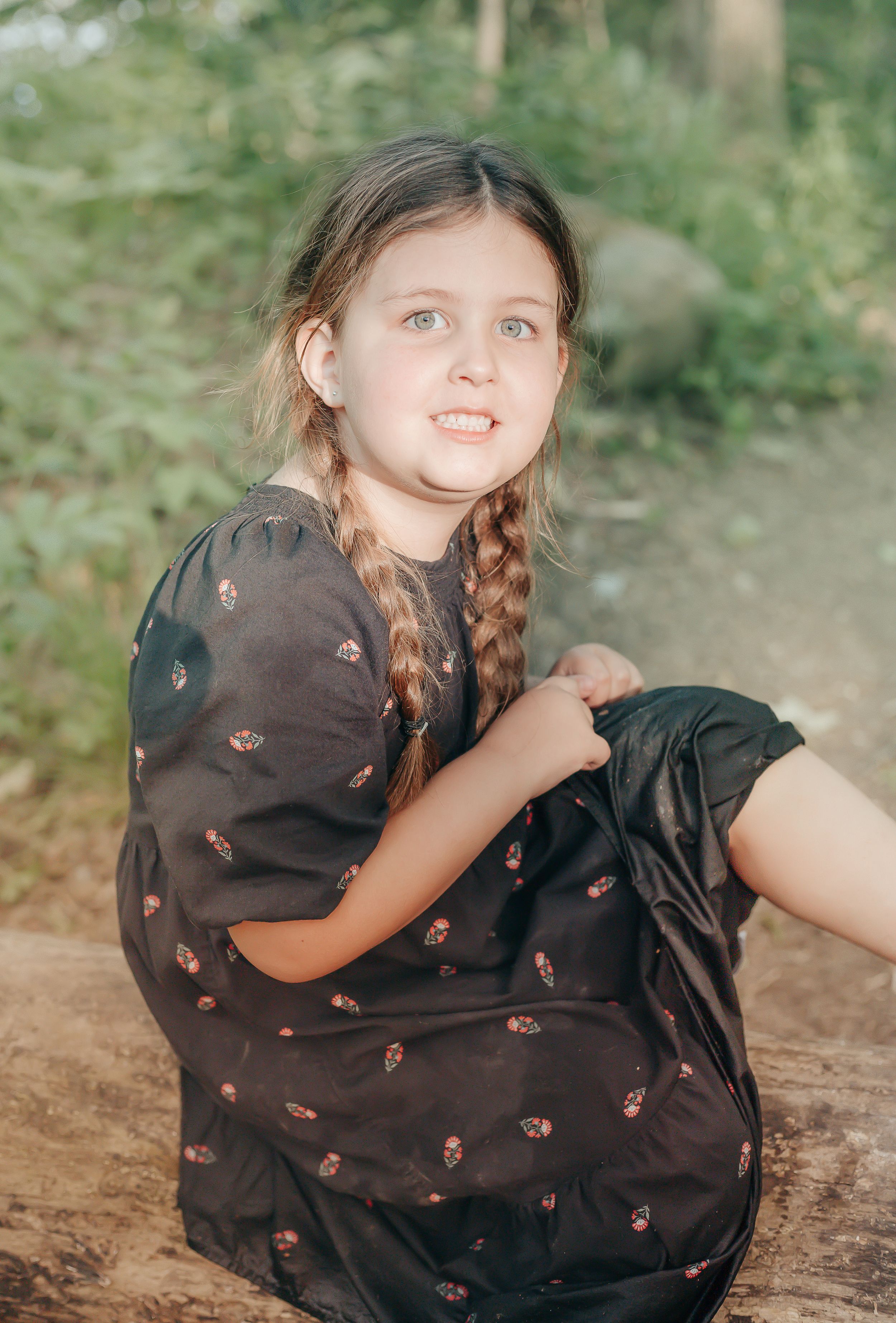 Young girl in dark floral dress sitting on rocky ground with braided hair.
