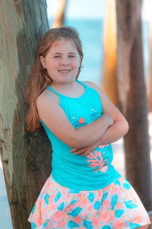 Girl in turquoise tank top with coral print leaning against wooden pier post.
