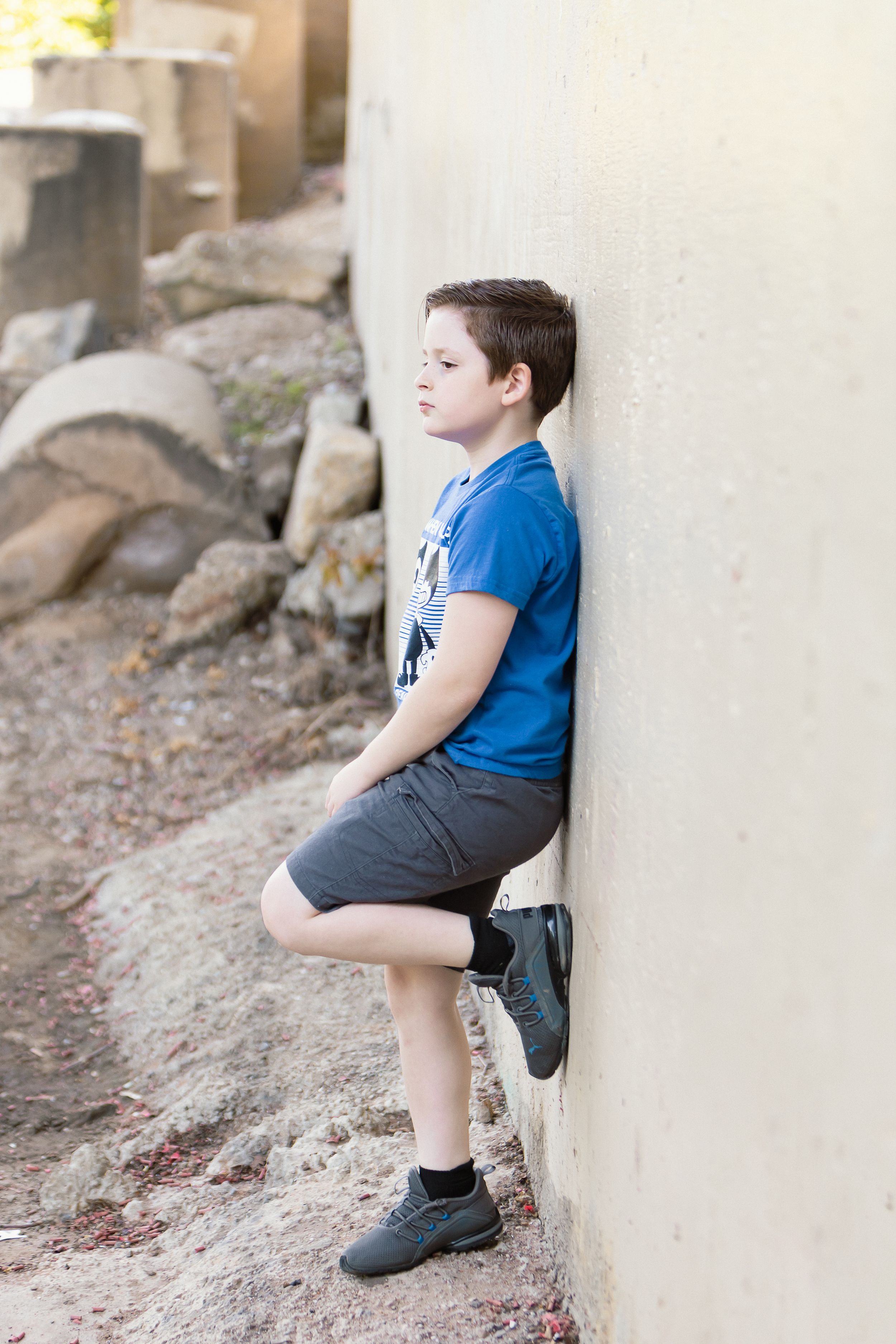 Boy in blue shirt leaning against beige wall with one foot propped up.