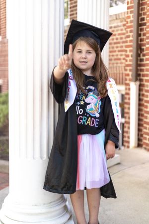 Graduate in cap and gown with unicorn shirt poses by white column.