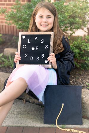 Graduate in cap and gown holds letter board next to diploma on brick steps.