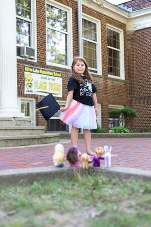 Standing in front of a brick school building with small dolls on the ground.