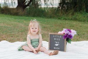 A toddler in mint green dress sits on white blanket with letterboard and purple flowers outdoors.