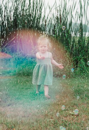 Small child in green dress chases bubbles in dreamy outdoor setting with tall grass.