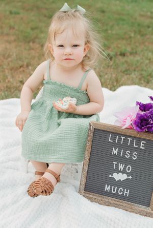 Small toddler in mint dress sits on white blanket with letterboard sign and purple flowers.