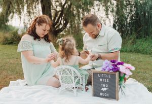Family sits on white blanket outdoors with decorative items and purple flowers.