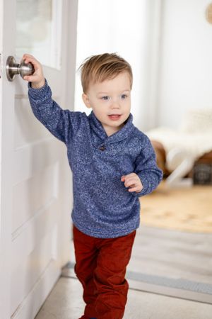 A toddler in a blue sweater and red pants reaching for a door handle in a bright room.