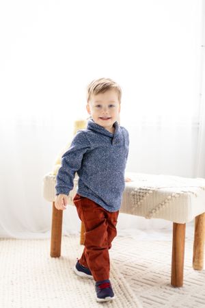 A young child in a navy pullover and red pants poses next to a wooden bench.