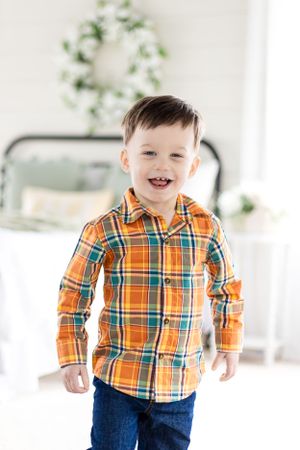 A cheerful child wearing a bright orange plaid shirt and jeans in a bright white room with greenery.