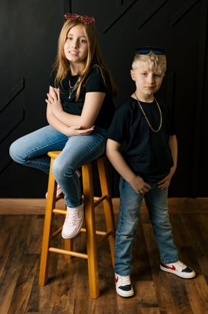 Two young people in matching black shirts pose together near a wooden stool.