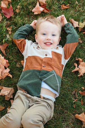 Small child lying on grass wearing color block jacket in earth tones surrounded by autumn leaves.