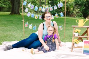 A picnic scene with a blanket on grass, displaying hanging photos with a yellow watering can and decorative setup.
